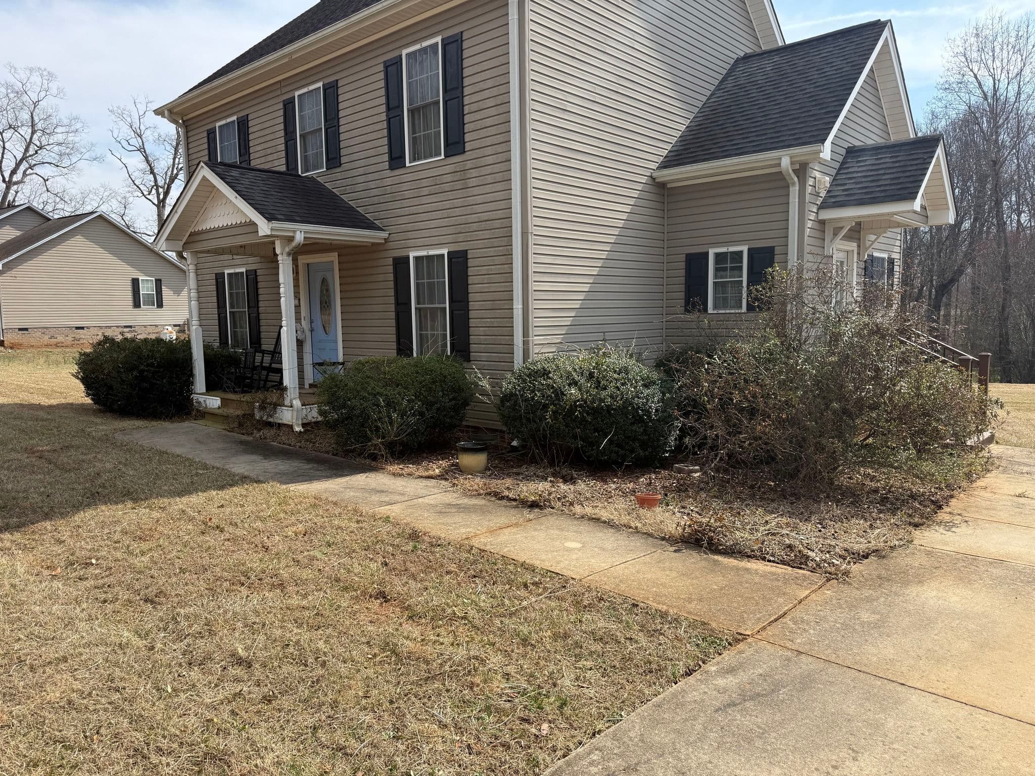 Two-story beige house with black shutters and a front porch, surrounded by grass and shrubs.