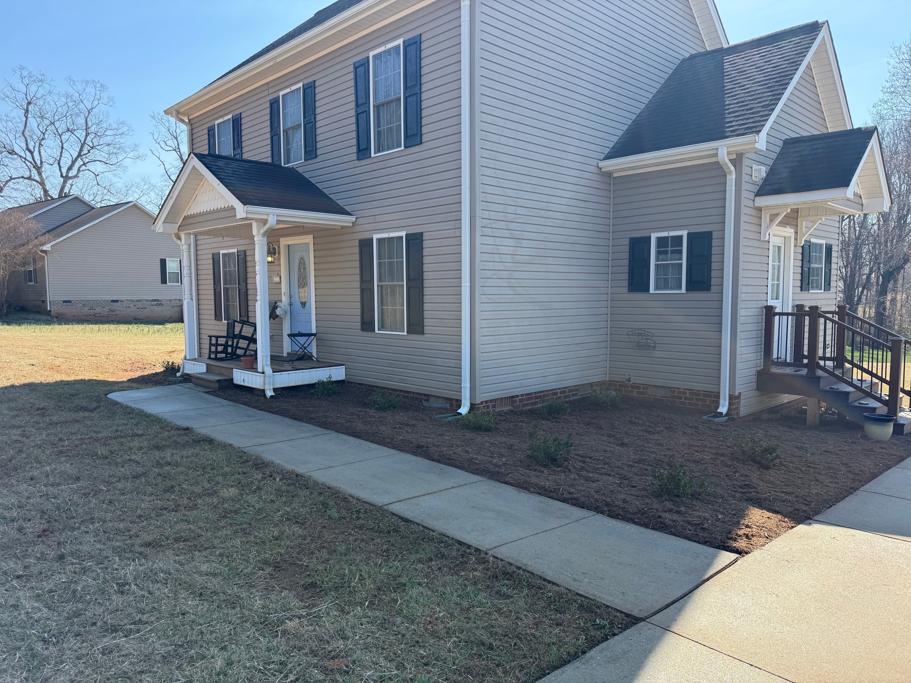 Two-story beige house with black shutters, front porch, and concrete walkway on sunny day.
